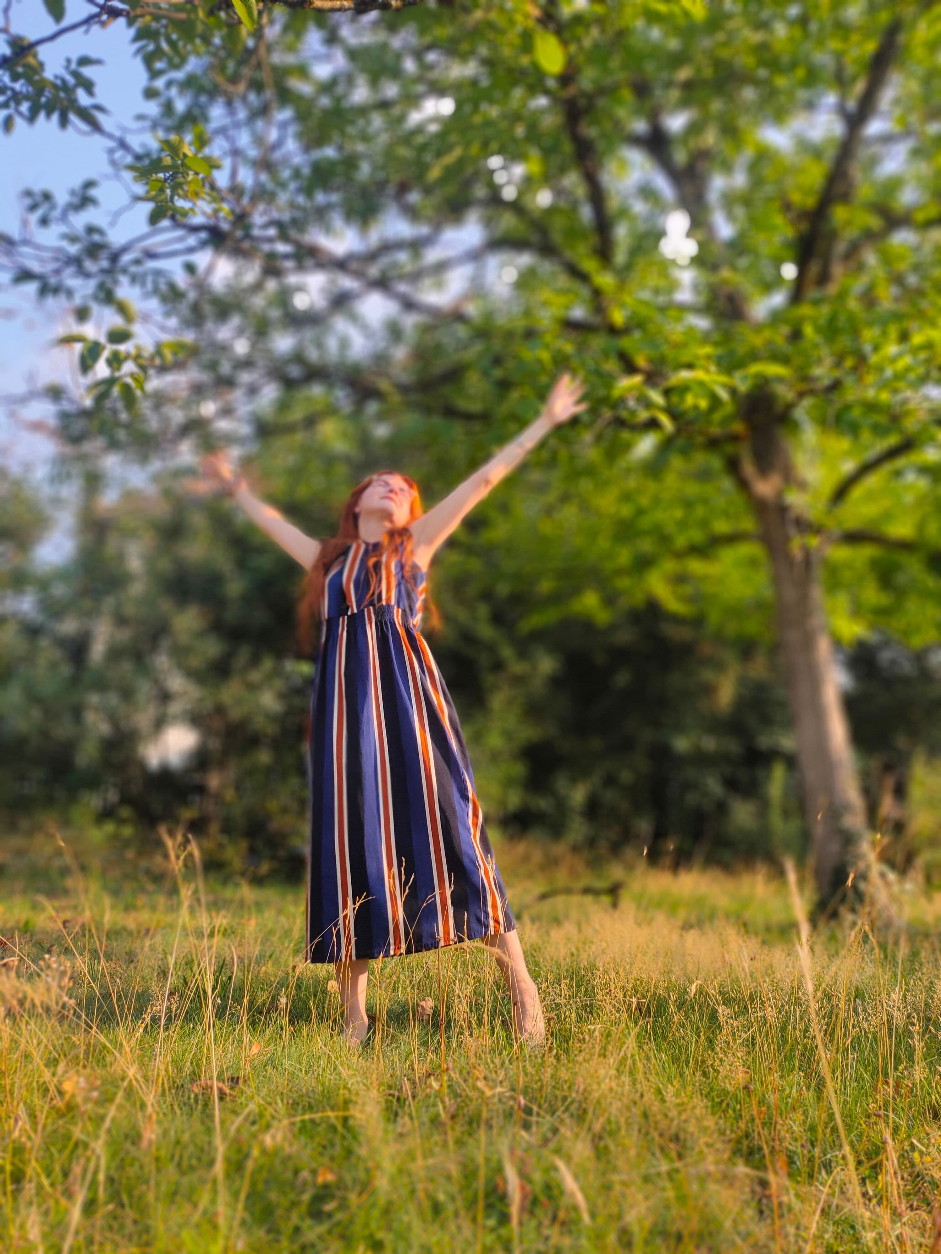 Luisa praktiziert eine fröhliche, vom Yoga inspirierte Dehnung im Stehen, barfuß auf einer sonnenbeschienenen Wiese, mit erhobenen Armen zum Himmel, umgeben von üppigen grünen Bäumen und der Natur.