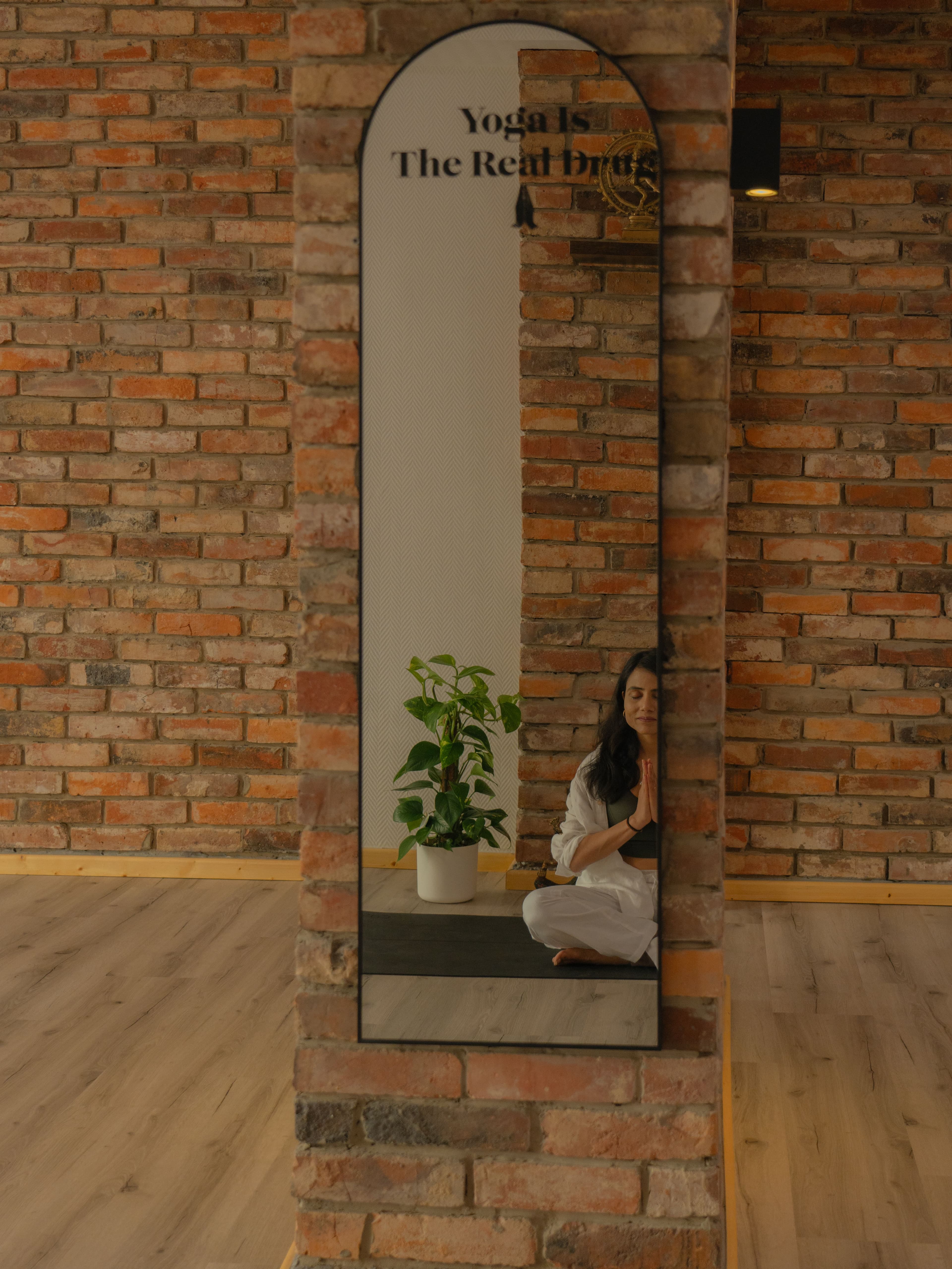 Bogenspiegel mit dem Schriftzug 'Yoga Is The Real Drug' an einer Backsteinsäule; im Spiegelbild meditiert eine Teilnehmerin im Yogicescape Friedrichshain Yoga Studio in Berlin